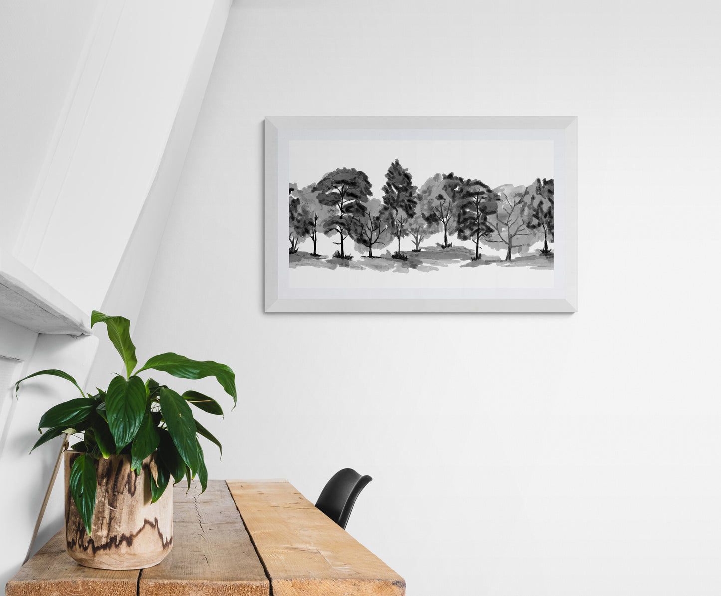 Framed black and white landscape of trees on a white wall above a wooden table with a plant.
