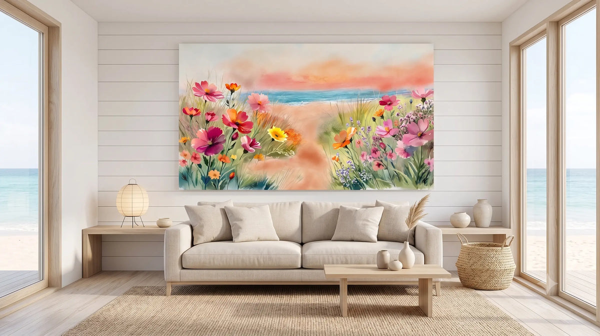 A large rectangular wall art print showing a beach and wildflowers hangs above a light beige sofa in a bright, modern living room with ocean views through tall windows.