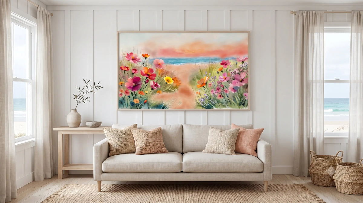 A framed colorful floral beach painting hangs on a white paneled wall above a light beige sofa in a bright, modern living room with windows, curtains, and beach decor.